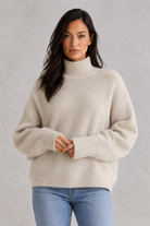Woman wearing a beige sweater and blue jeans against a gray background