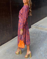 Woman in a patterned dress holding an orange clutch against a black wall.