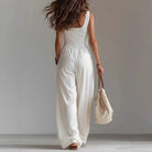 Woman wearing a white sleeveless jumpsuit holding a beige bag against a plain background