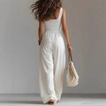 Woman wearing a white sleeveless jumpsuit holding a beige bag against a plain background