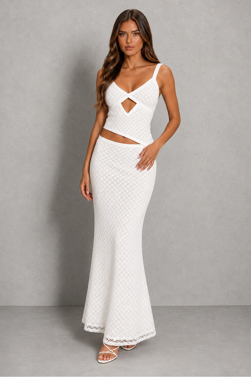 A woman modeling a white two-piece knit maxi dress set against a neutral gray studio backdrop. The dress features a sleeveless, textured crop top with a crossover sweetheart neckline and a matching high-waisted, flared maxi skirt.