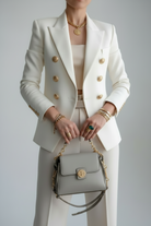 Person wearing a white suit with gold buttons holding a gray handbag against a plain background