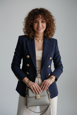 Person wearing a navy blazer with gold buttons, holding a gray handbag with gold chain strap.