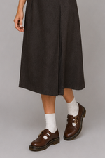 Woman wearing a brown shirt, black skirt, and brown shoes on a gray background