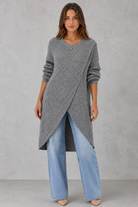 Woman wearing a long gray sweater with an asymmetrical hem and light blue jeans on a gray background