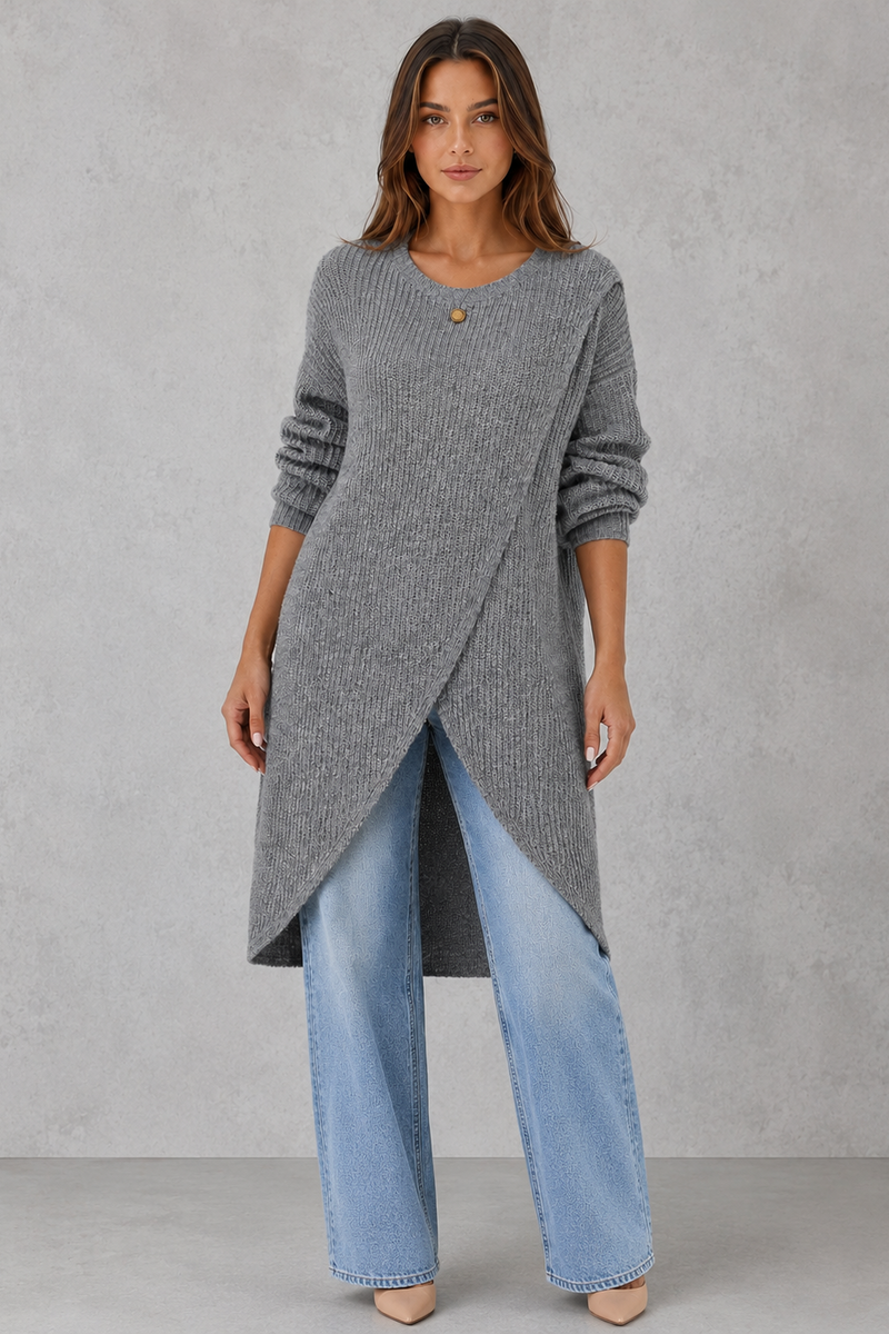 Woman wearing a long gray sweater with an asymmetrical hem and light blue jeans on a gray background