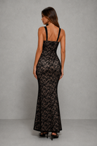 A woman modeling a black lace maxi dress from the back, featuring thin shoulder straps, a fitted silhouette, and a flared hem, set against a neutral gray studio background.