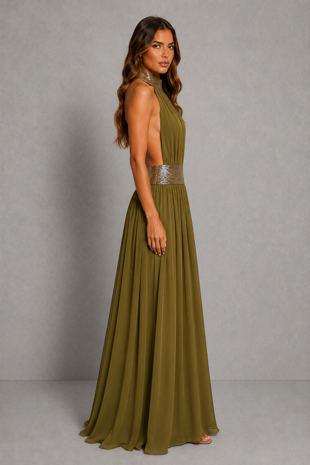 A woman modeling a sleeveless olive green maxi dress with a halter neckline, deep V-back, and a metallic silver embellished waistband, set against a neutral gray studio backdrop.