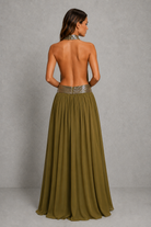 A woman shown from the back wearing a long, open-back olive green dress with a metallic silver patterned waistband, set against a textured gray studio background.