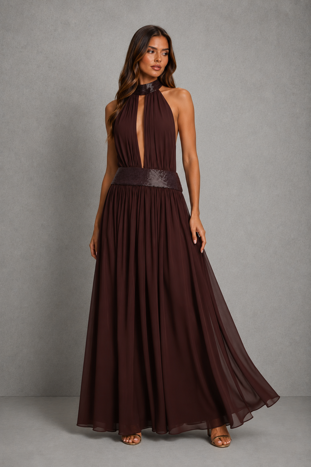 A woman modeling a floor-length halter gown in deep chocolate brown. The dress features a structured, sheer-like bodice with a plunging V-neckline, a wide matching waistband, and a flowing chiffon-like skirt, set against a neutral gray studio backdrop.