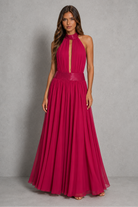A woman modeling a sleeveless, high-neck, floor-length magenta pink gown with a pleated skirt and a matching waistband, set against a neutral gray studio backdrop.