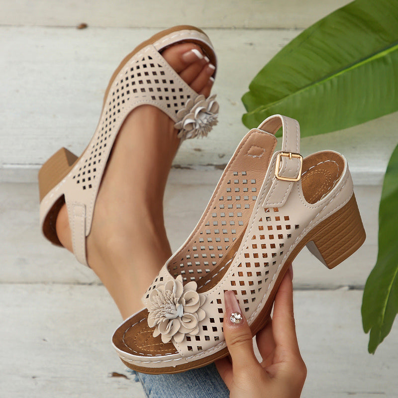 Beige perforated sandals with floral details worn by a person on a light wooden floor.