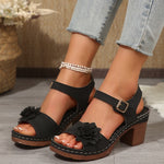 Black sandals with floral details worn by a person on a wooden floor.