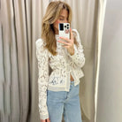 Woman taking a mirror selfie wearing a white lace top and blue jeans against a beige curtain.