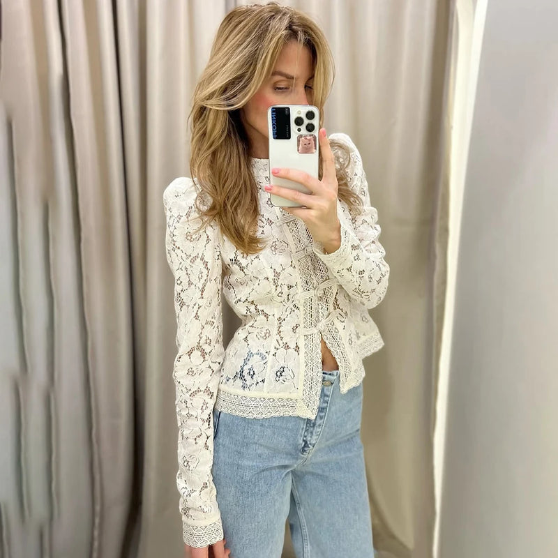 Woman taking a mirror selfie wearing a white lace top and blue jeans against a beige curtain.