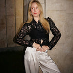 Woman wearing a black lace top and white pants against a textured wall.