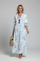 Woman wearing a long, light blue dress with white patterns, standing against a white wall.