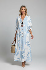 Woman wearing a long, light blue dress with white patterns, standing against a white wall.