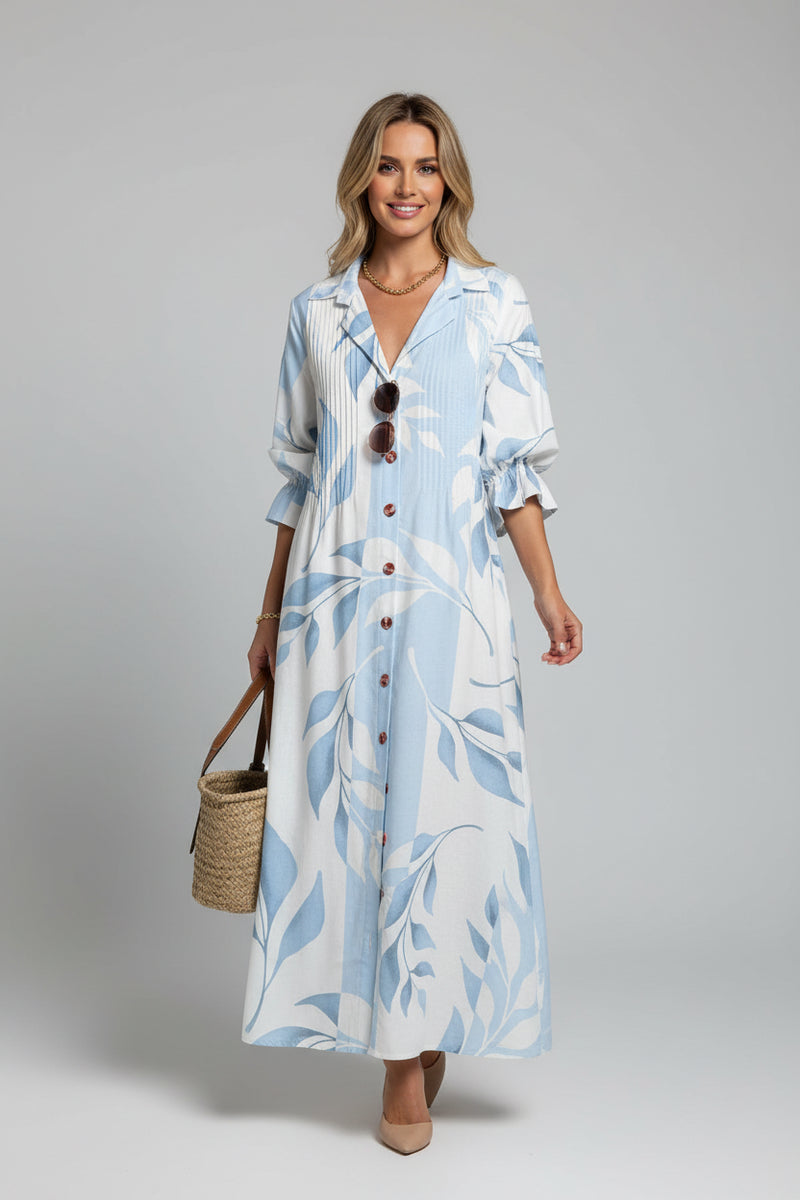 Woman wearing a long, light blue dress with white patterns, standing against a white wall.
