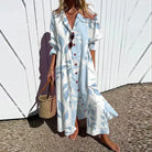 Woman wearing a long, light blue dress with white patterns, standing against a white wall.