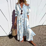 Woman wearing a long, light blue dress with white patterns, standing against a white wall.