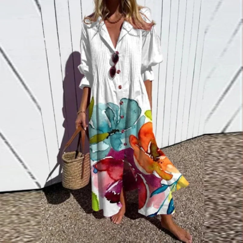 Woman wearing a colorful floral dress with a white shirt over it, standing against a white wall.