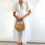 Woman wearing a white dress holding a woven handbag against a plain background