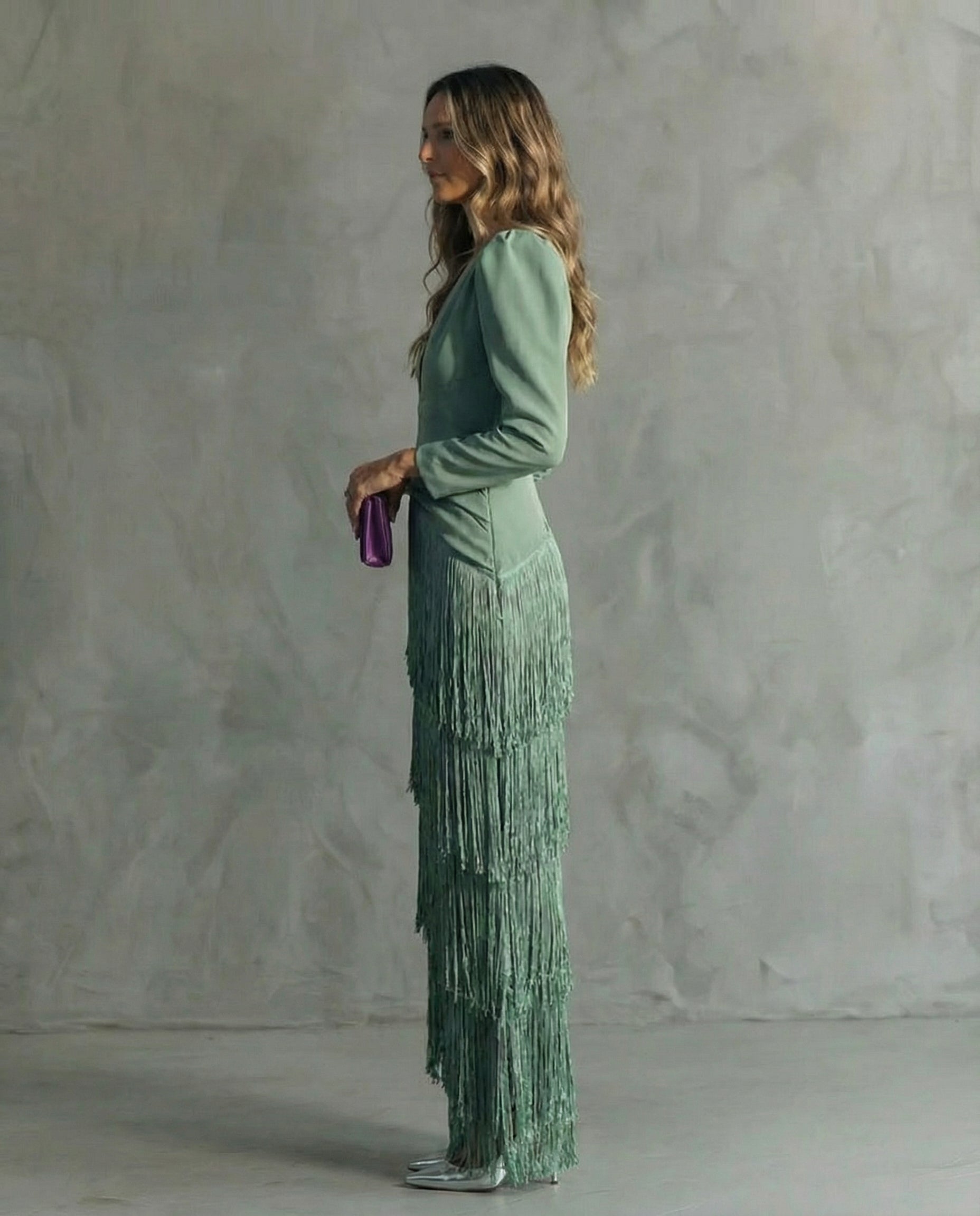 Woman in a green fringe dress holding a purple clutch against a gray background