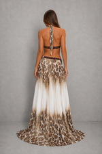 A woman shown from the back wearing a sleeveless, open-back maxi dress with a leopard print pattern that fades into a white gradient at the hem, set against a neutral gray studio background.