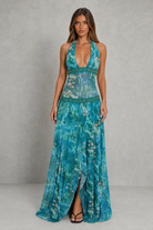 A woman modeling a sleeveless halter-neck maxi dress with a deep plunging neckline, a teal and blue tropical floral print, and a high slit, set against a gray studio backdrop.