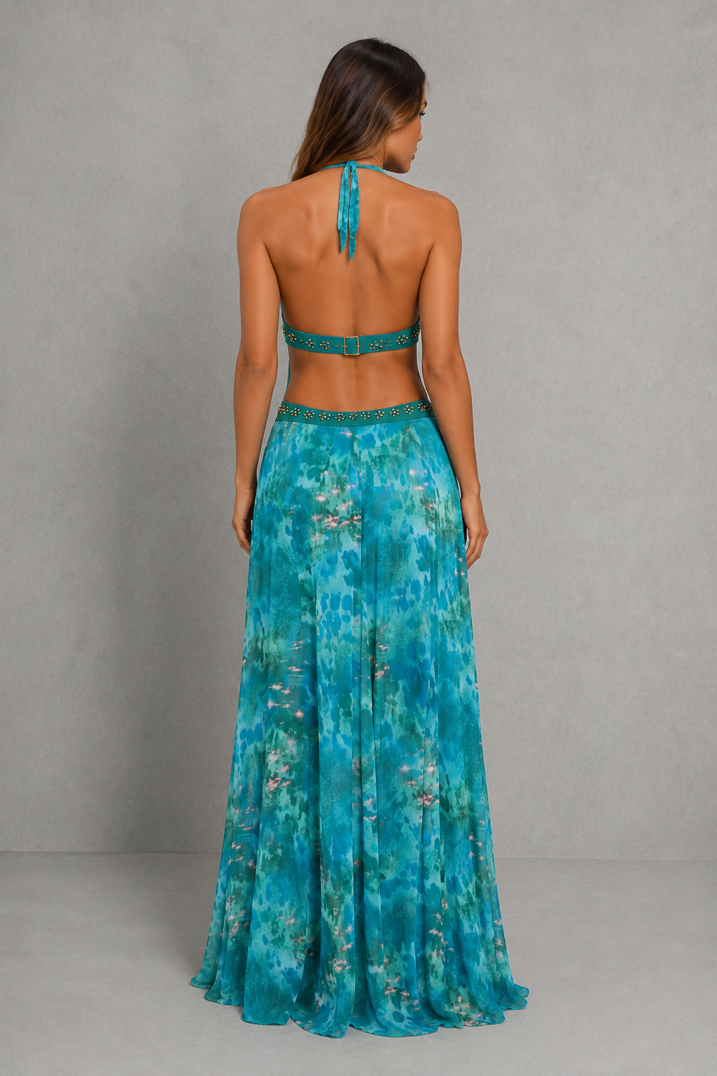 A woman viewed from the back wearing a teal and turquoise floral halter-neck maxi dress with a matching embellished waistband, set against a neutral gray studio backdrop.