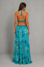 A woman viewed from the back wearing a teal and turquoise floral halter-neck maxi dress with a matching embellished waistband, set against a neutral gray studio backdrop.