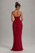 A woman shown from the back wearing a floor-length red gown with a halter neckline, structured corset-style back, and dramatic gathered draping on the skirt, photographed against a plain light gray studio backdrop.