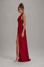 A woman modeling a floor-length, sleeveless red gown with a high thigh slit, photographed against a neutral gray studio backdrop.