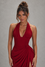 A woman modeling a deep red halter-neck dress with a plunging V-neckline, ruched bodice, and draped skirt details against a neutral gray studio background.