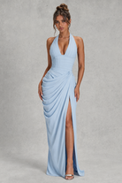 A woman modeling a light blue halter-neck maxi dress with a deep V-neckline, draped gathered skirt, and high front slit, standing against a textured gray studio backdrop.