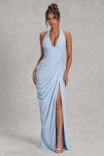 A woman modeling a light blue halter-neck maxi dress with a deep V-neckline, draped gathered skirt, and high front slit, standing against a textured gray studio backdrop.