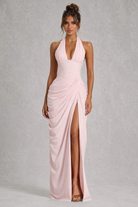 A woman modeling a light pink halter-neck maxi dress with a draped, gathered skirt and a high thigh slit, set against a textured gray studio background.