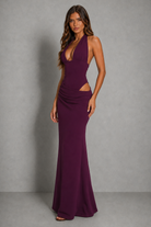 A woman modeling a sleeveless, deep V-neck floor-length gown in a rich plum purple color, featuring a halter-style neckline, ruched bodice, and side cutouts, set against a neutral gray studio backdrop.