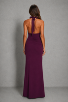 A woman viewed from the back wearing a sleeveless, open-back floor-length gown in deep burgundy with a tie at the neck, standing against a plain gray studio backdrop.