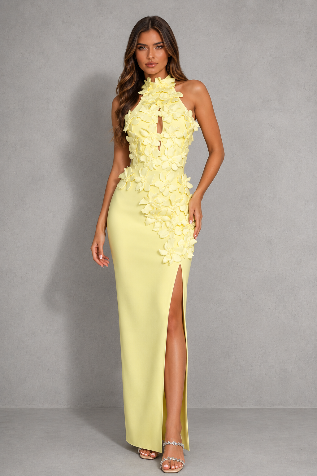 A woman modeling a pale yellow sleeveless maxi dress with 3D floral applique on the bodice and hips, a high slit, and a keyhole cutout at the neckline, set against a gray studio backdrop.