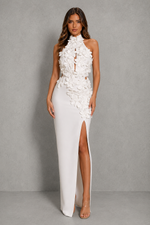 A woman modeling a sleeveless white halter-neck maxi gown with a high thigh slit and 3D floral applique on the bodice and hip, set against a textured gray studio backdrop.