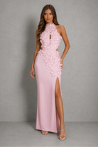 A woman modeling a sleeveless, high-neck, floor-length pink gown with 3D floral applique on the bodice and hips, set against a neutral gray studio backdrop.