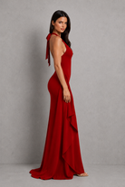 A woman modeling a floor-length red halter gown with a side tie detail and layered ruffle skirt, photographed against a neutral gray studio backdrop.