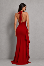 A woman wearing a floor-length red evening gown, shown from the back against a textured gray studio backdrop. The dress has a halter-style open back with a large bow, a fitted silhouette, and layered ruffle draping along the side.