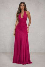 A woman modeling a floor-length magenta halter gown with a deep V-neckline, ruched bodice, and flowing pleated skirt against a textured gray studio backdrop.