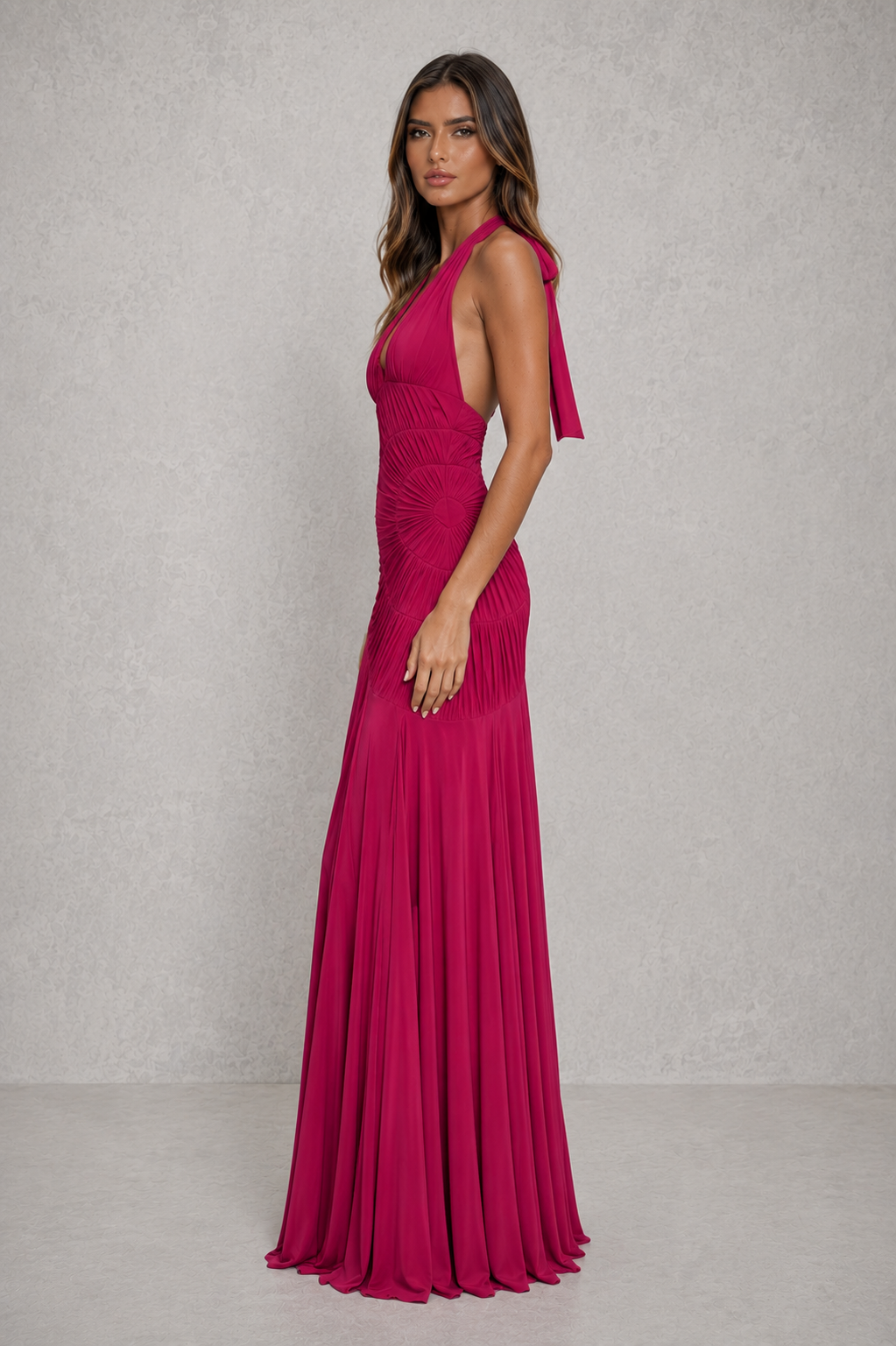 A woman modeling a sleeveless, high-neck, floor-length magenta gown with a pleated skirt, standing against a textured light gray studio backdrop.