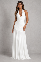A woman modeling a floor-length white halter gown with a plunging V-neckline, gathered corset-style bodice, and flowing skirt against a textured gray studio backdrop.