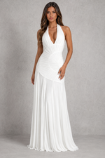 A woman modeling a floor-length white halter gown with a plunging V-neckline, gathered corset-style bodice, and flowing skirt against a textured gray studio backdrop.