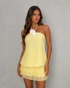 A woman modeling a pale yellow halter-neck mini dress with a ruffled tiered hem and a large white flower at the neckline, set against a neutral gray studio background.
